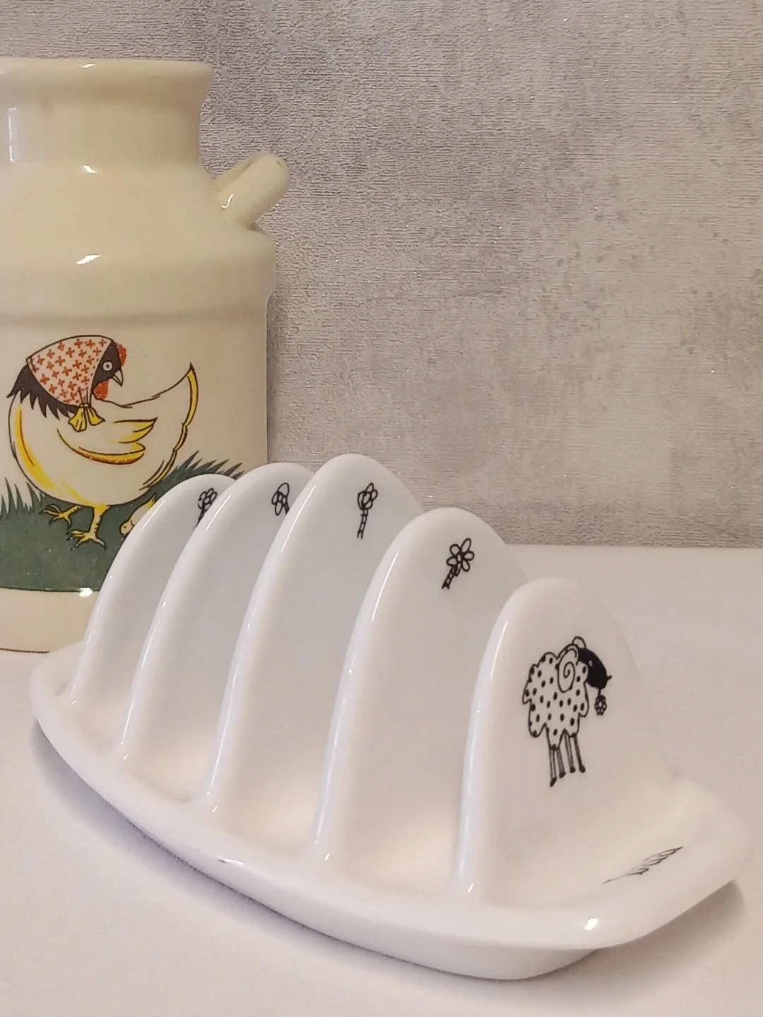 Arka planda tavuk desenli süt güğümü bulunan, bölmelerinde siyah çizgi koyun ve çiçek figürleri olan beyaz toast rack
