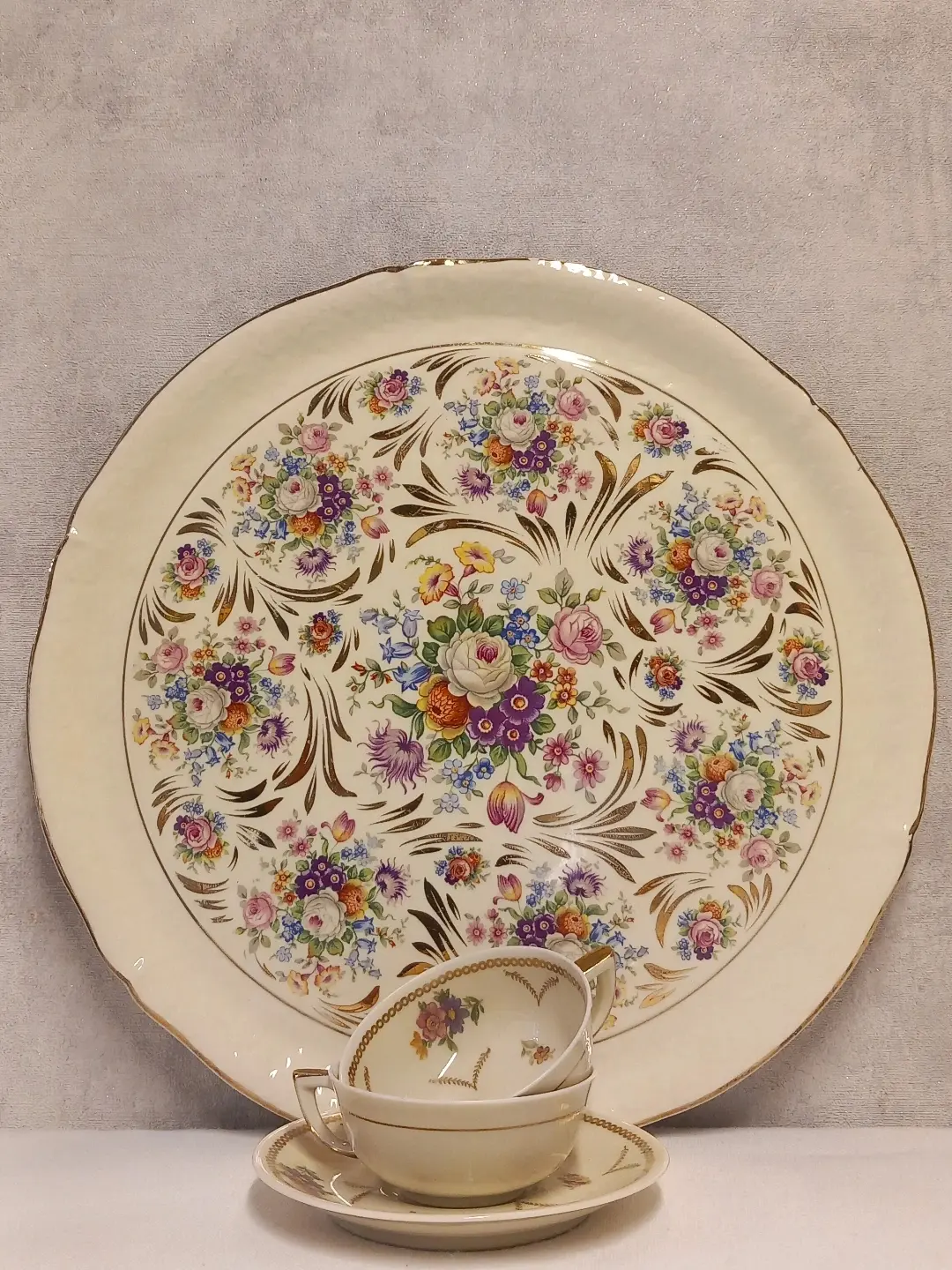 Mor ve pembe çiçek buketli, altın yaldız kıvrımlı krem Ceranord servis tabağı; önünde çiçekli fincan ve fincan tabakları