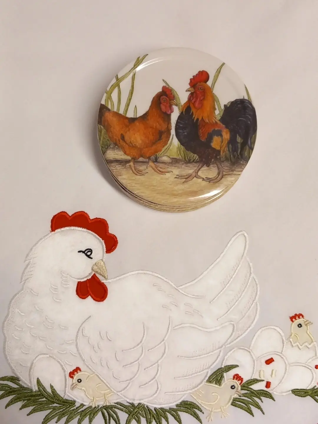 Kızıl tavuk ve siyah-turuncu horoz desenli istiflenmiş melamin bardak altlıkları; altta tavuk işlemeli örtü