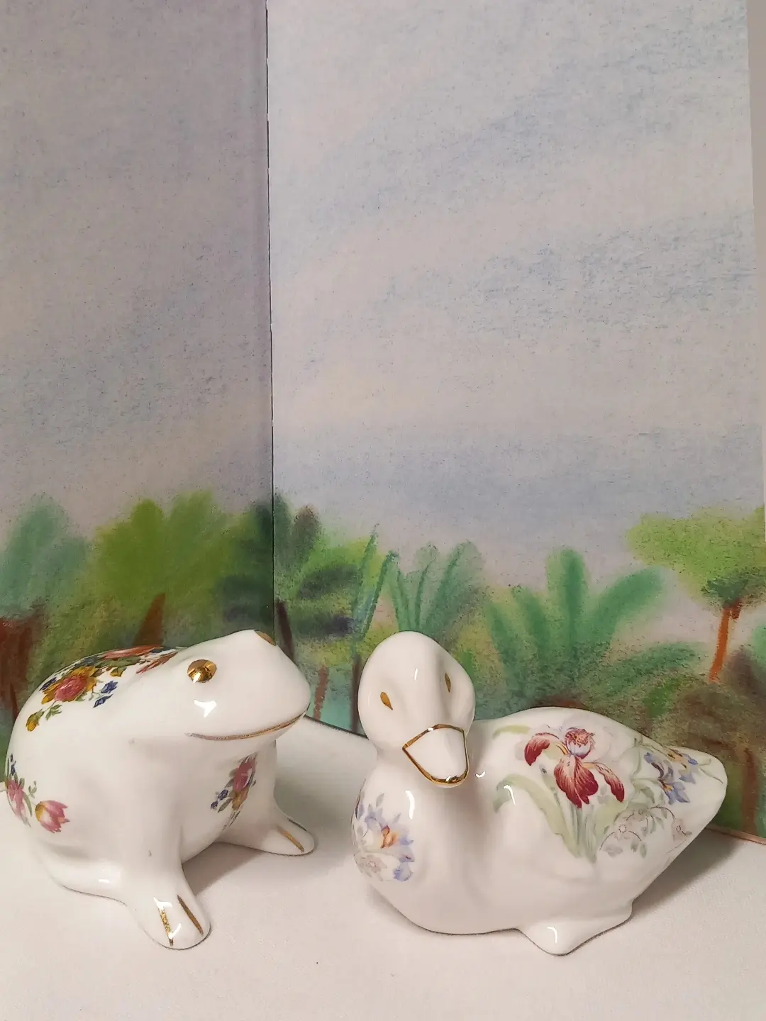 Gül desenli beyaz porselen kurbağa ve süsen desenli porselen ördek figürü; arkada pastel orman resmi
