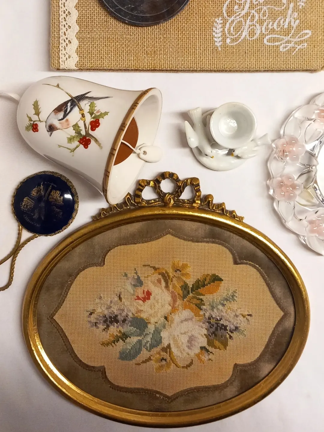 Oval yaldızlı çerçeveli goblen çiçek panosu; çevresinde kuş desenli çan, güvercinli mumluk ve cam tabak gibi başka objeler