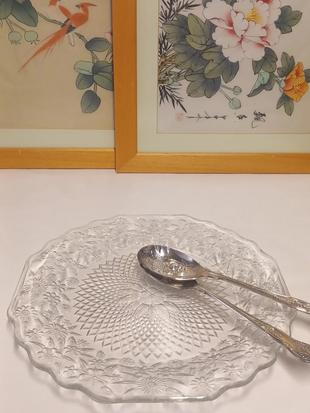 Çiçek ve kuş resimli çerçeveler önünde, üzerinde iki gümüş servis kaşığı duran kabartma desenli şeffaf cam tabak
